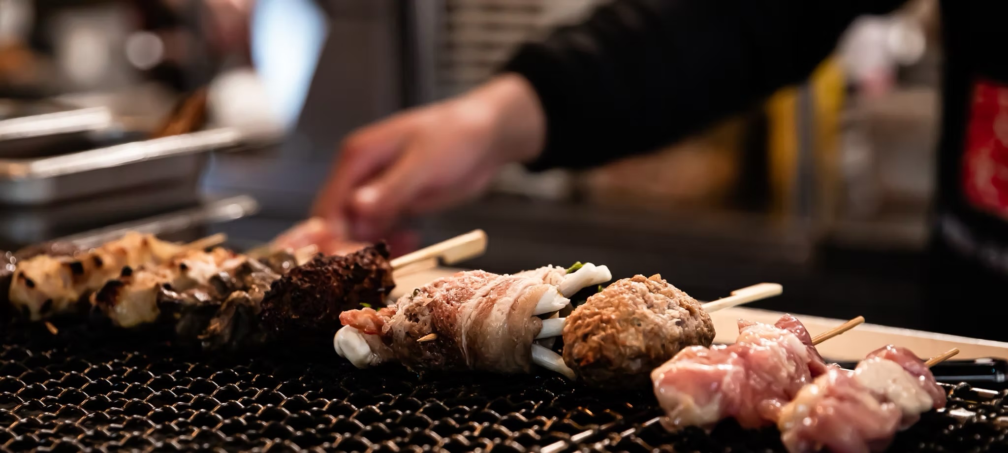 Chef's hand turning skewers over the charcoal grill, flames glowing beneath