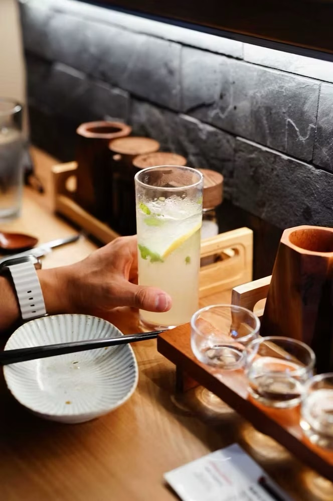 Hand lifting a yuzu highball at the counter beside a sake tasting flight