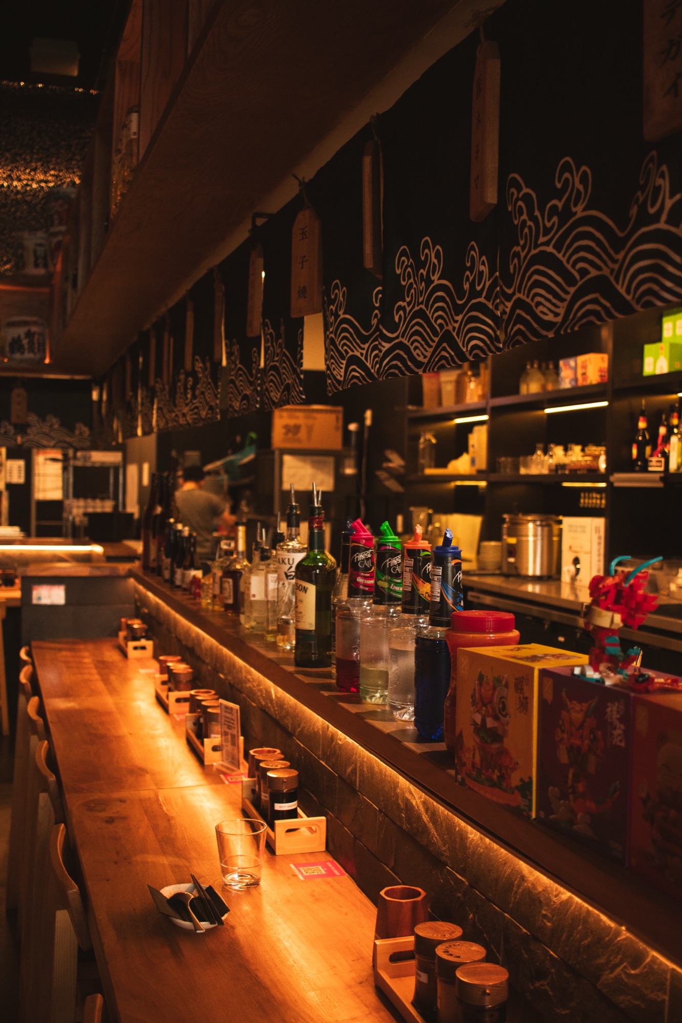 The bar at night, warm light on the bottles and the counter stretching back into the dark